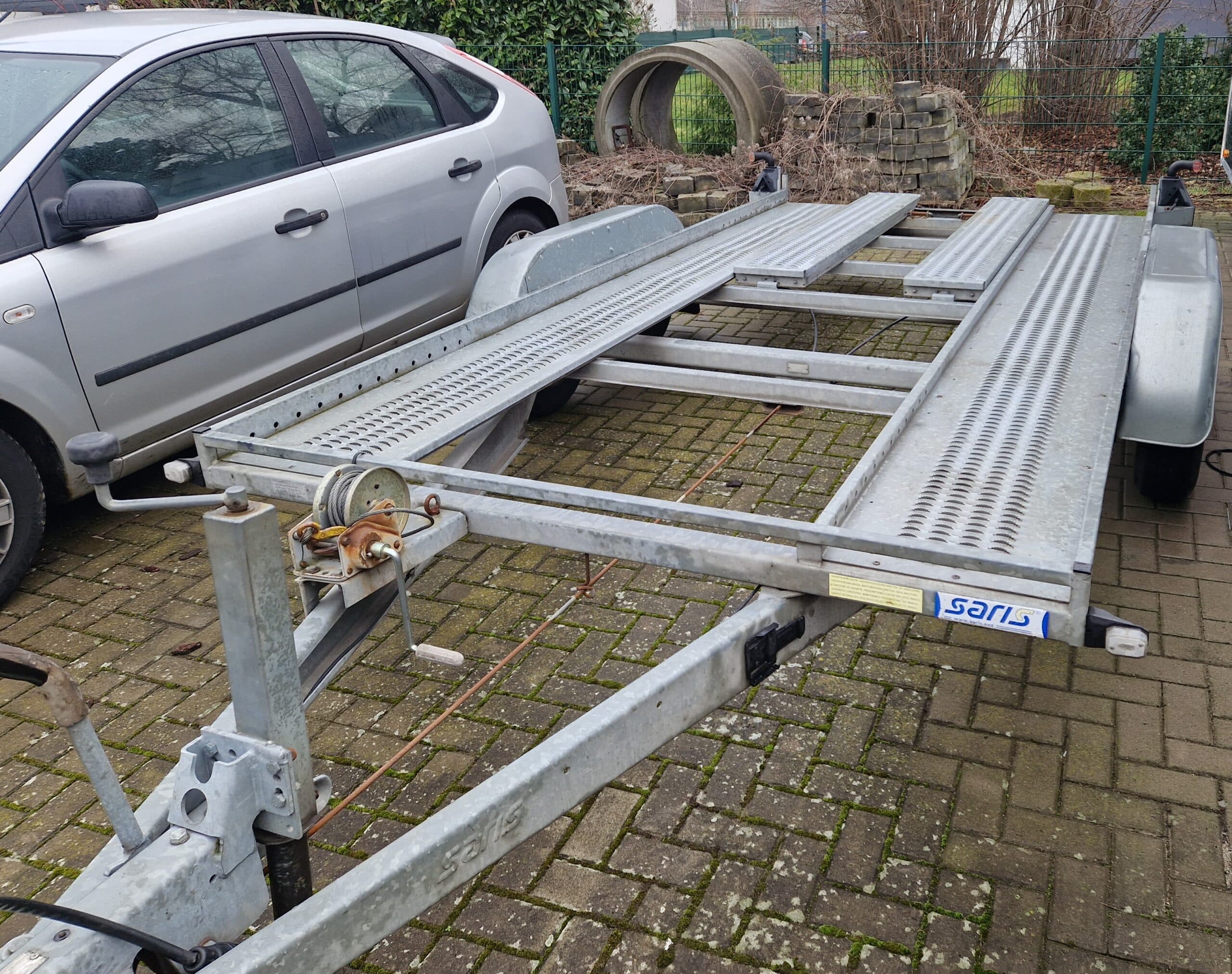 Ein Autoanhänger aus Metall mit Rampen ist auf einer gemauerten Auffahrt neben einem silbernen Auto geparkt. Der Anhänger hat eine Handwinde an der Vorderseite und gelochte Schienen für die Fahrzeugräder. Im Hintergrund sind ein Zaun und einige Rohre zu sehen.