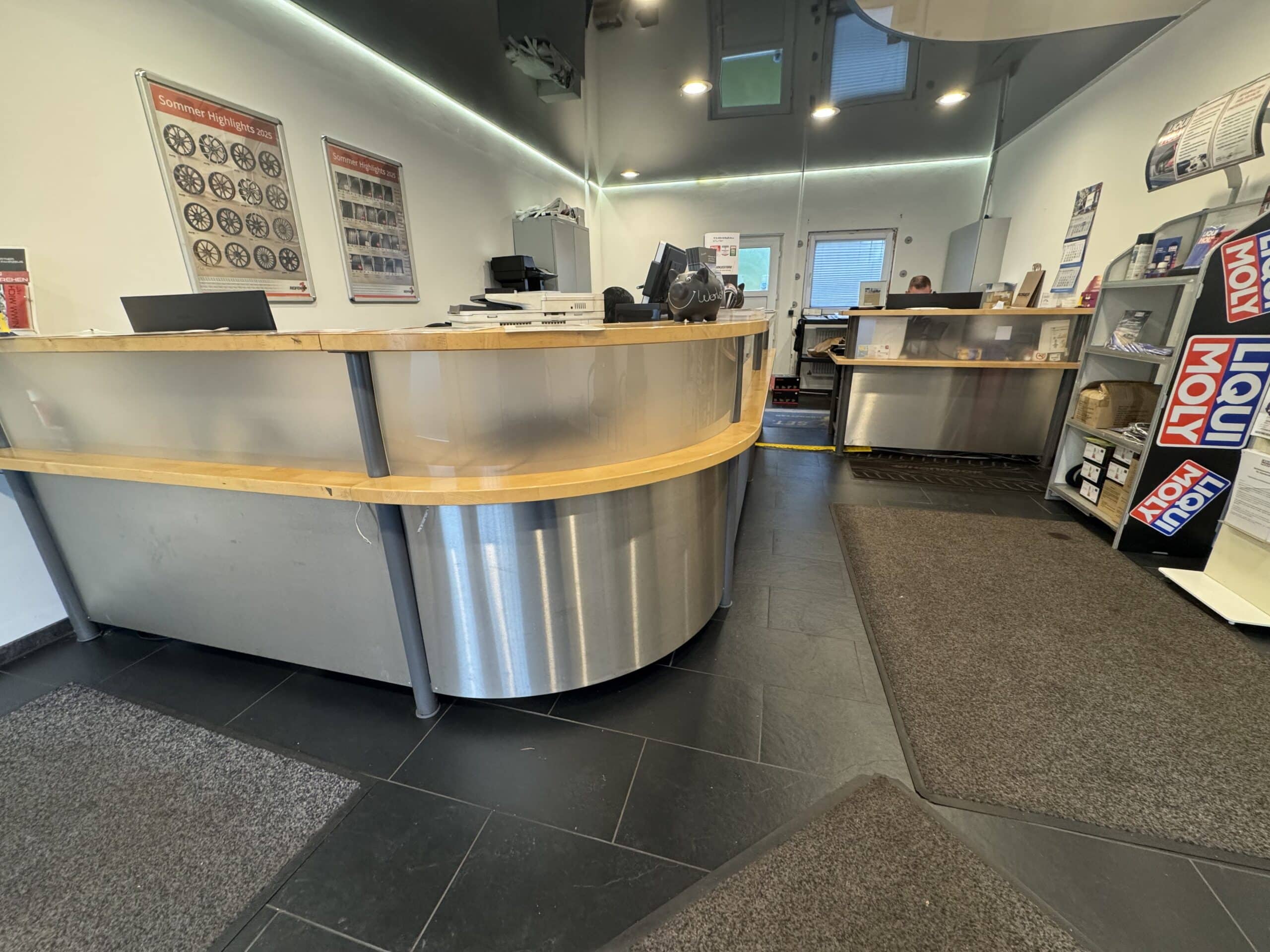 Ein Empfangsbereich mit einem geschwungenen Metall- und Holztresen, Büromaterialien, einem Computer, Regalen mit Broschüren über den Reifenservice, Wandpostern und Teppichböden auf schwarzem Fliesenbelag.