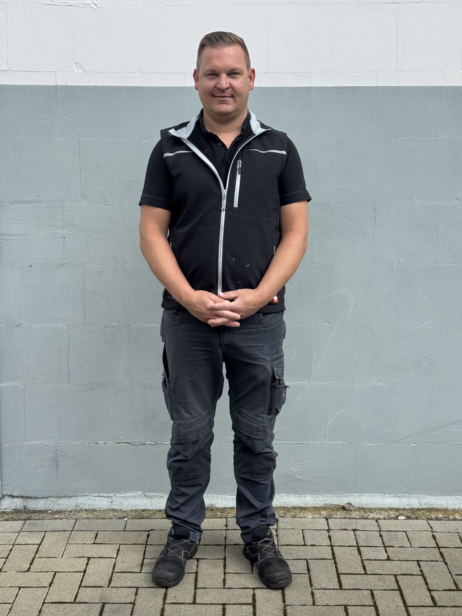 Ein Mann steht auf einer gepflasterten Fläche vor einer hellgrau-weißen Wand. Er trägt ein schwarzes, kurzärmeliges Hemd, eine dunkle Cargohose und schwarze Schuhe - bereit für den nächsten Reifendienst oder als Helfer beim Reifenwechsel. Seine Hände sind vor ihm verschränkt.