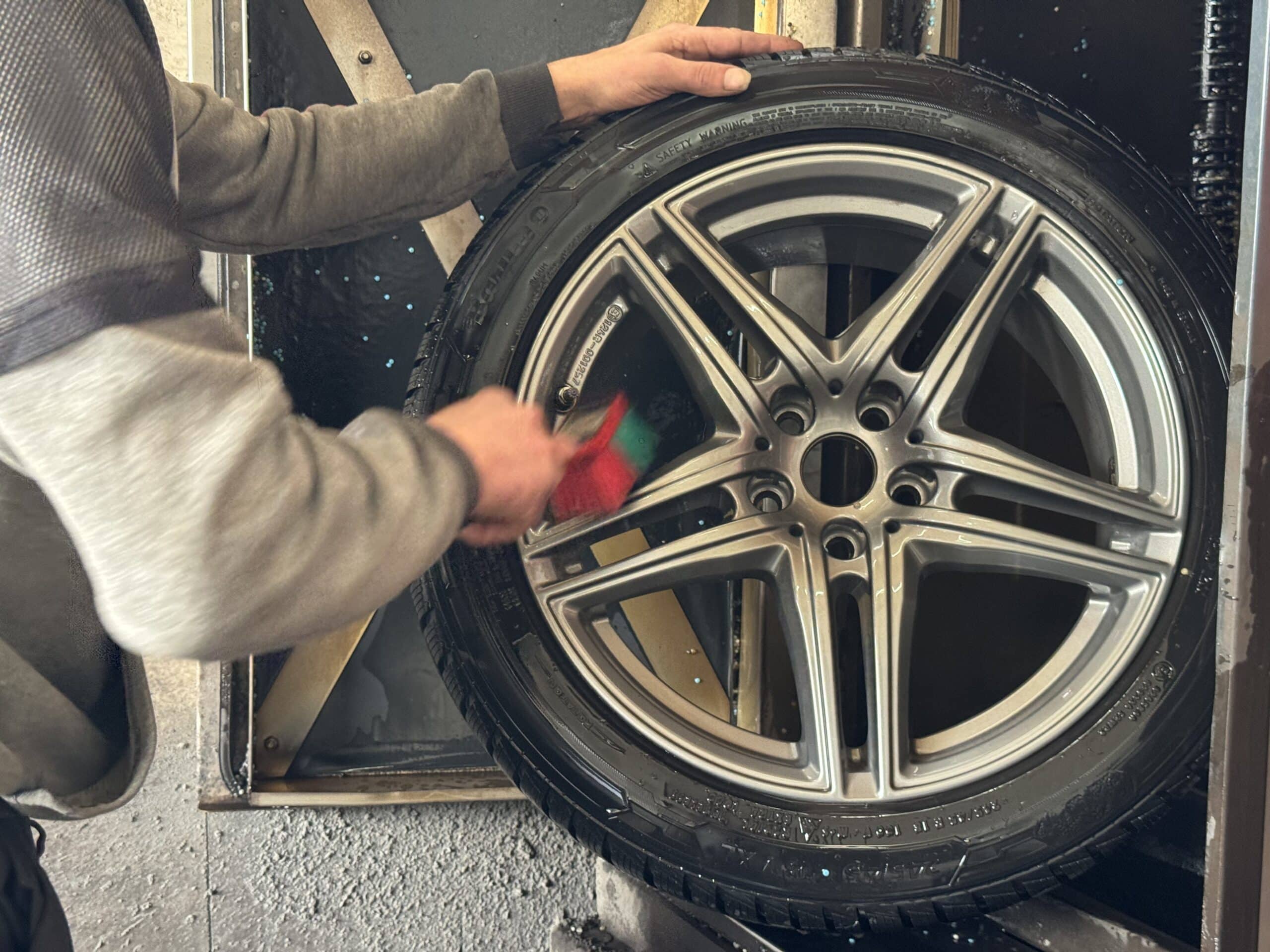 Eine Person verwendet eine Bürste, um die Speichen eines Autorads vor dem Reifenwechsel zu reinigen. Das Rad ist senkrecht montiert und scheint sich in einer Werkstatt oder Garage zu befinden, möglicherweise bei Reifen Wolfenbüttel.