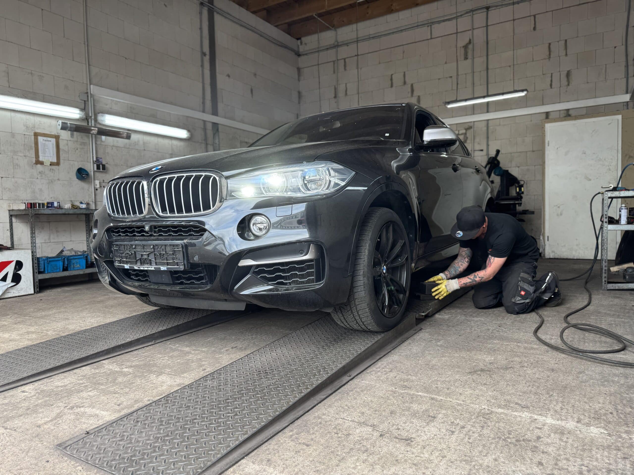 Ein Mechaniker mit Mütze und Handschuhen kniet neben einem schwarzen BMW-Geländewagen und führt einen Reifenservice am rechten Vorderrad in einer Werkstatt mit weißen Backsteinwänden und Deckenbeleuchtung durch.