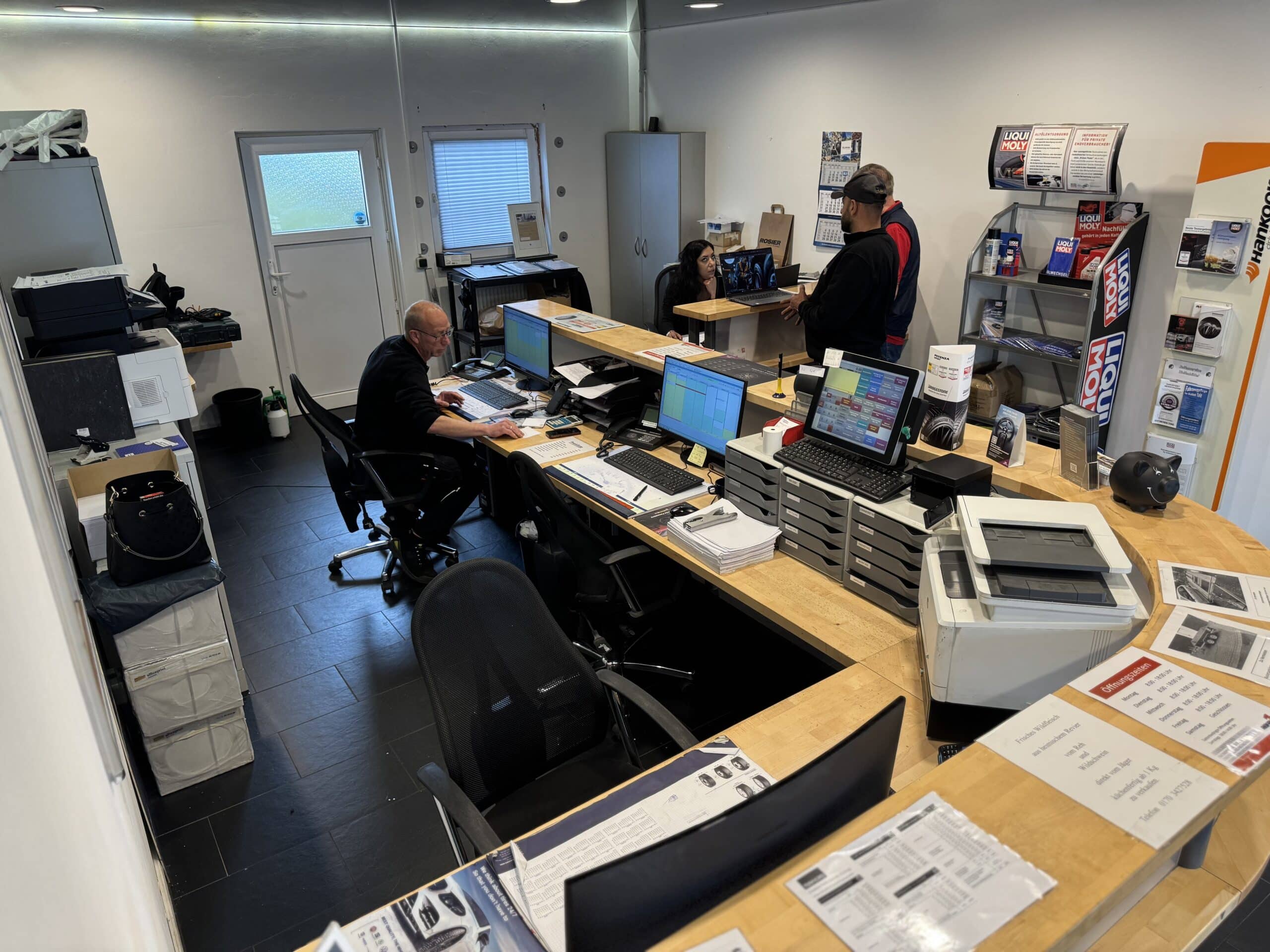 Empfangsbereich des Büros mit drei Personen: eine arbeitet am Schreibtisch, eine weitere steht am Tresen und eine dritte sitzt gegenüber. In den Regalen sind Produkte und Büroausstattung ausgestellt, die die professionelle Reifenservice-Atmosphäre von Reifen Wolfenbüttel unterstreichen.