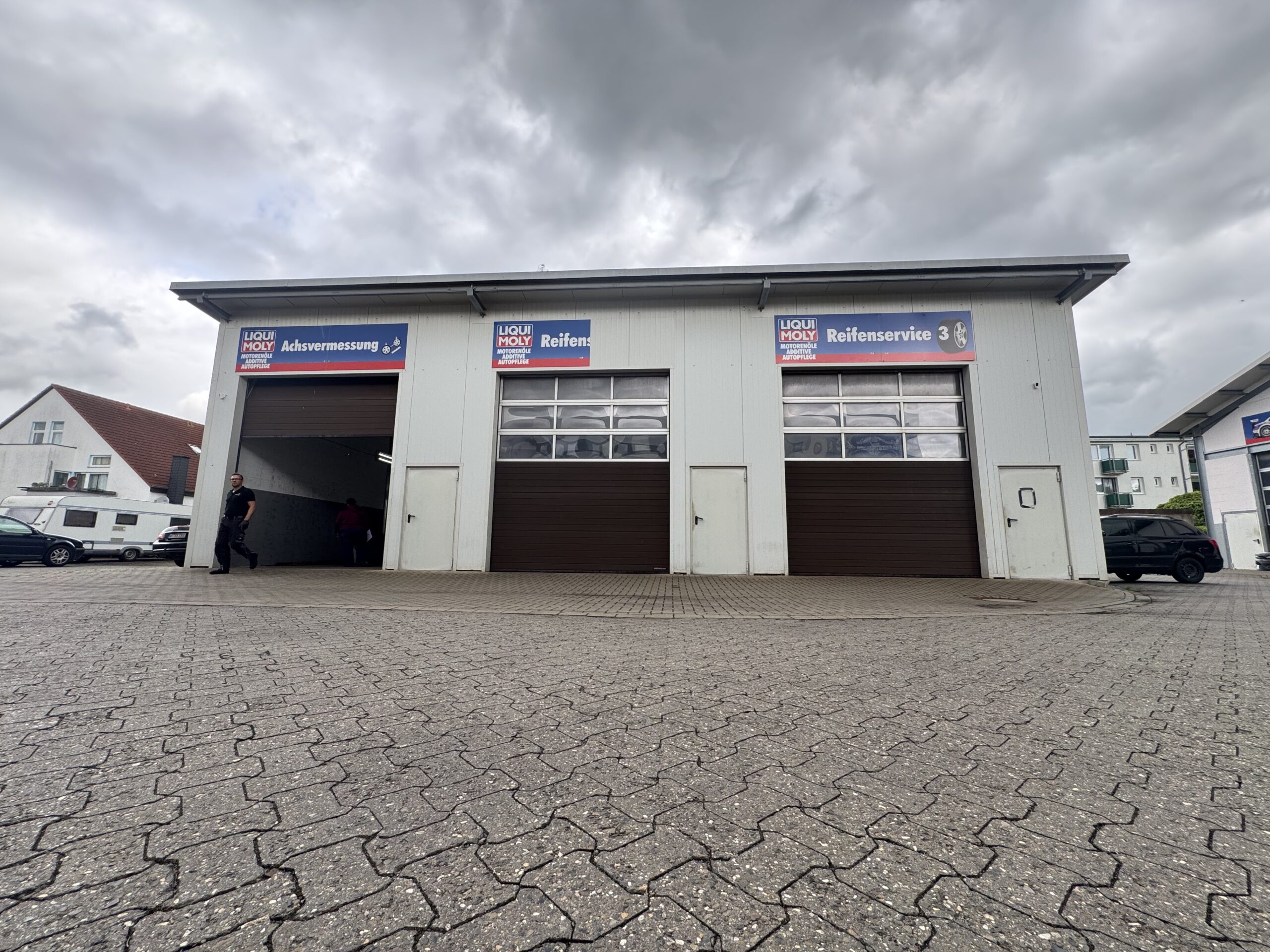 Blick aus geringer Höhe auf eine Autowerkstatt mit drei großen Toren, zwei davon geschlossen. Auf den Schildern in deutscher Sprache stehen "Reifenservice" und "Reifen Wolfenbüttel". Das Gebäude ist von einer gepflasterten Fläche unter bewölktem Himmel umgeben.