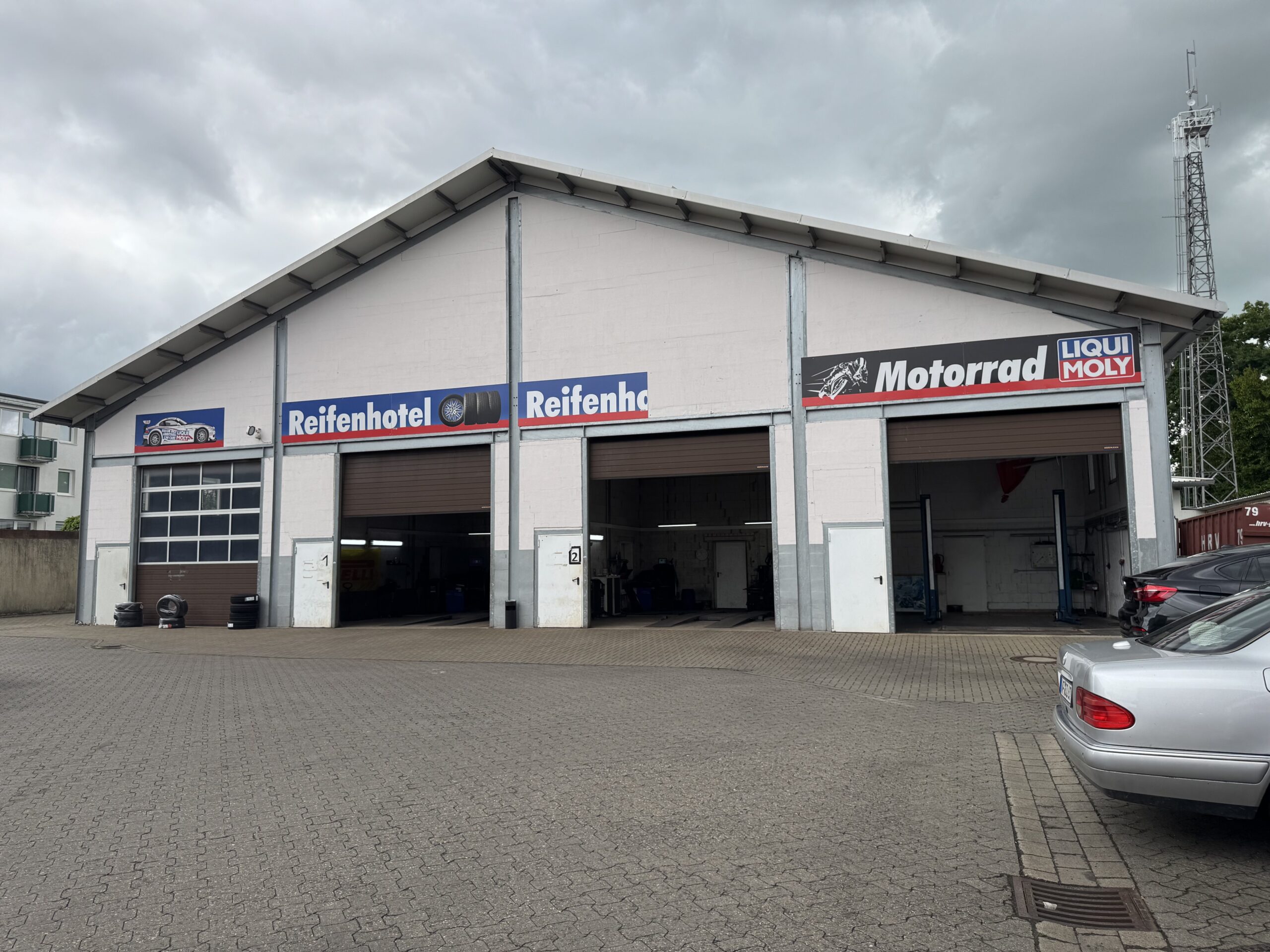 Eine große weiße Autowerkstatt in Wolfenbüttel mit drei offenen Buchten bietet Reifenservice und Reifenwechsel an. Auf den Schildern darüber steht "Reifenhotel", "Reifenhotel" und "Motorrad LIQUI MOLY". Mehrere Autos sind unter dem wolkenverhangenen Himmel geparkt.