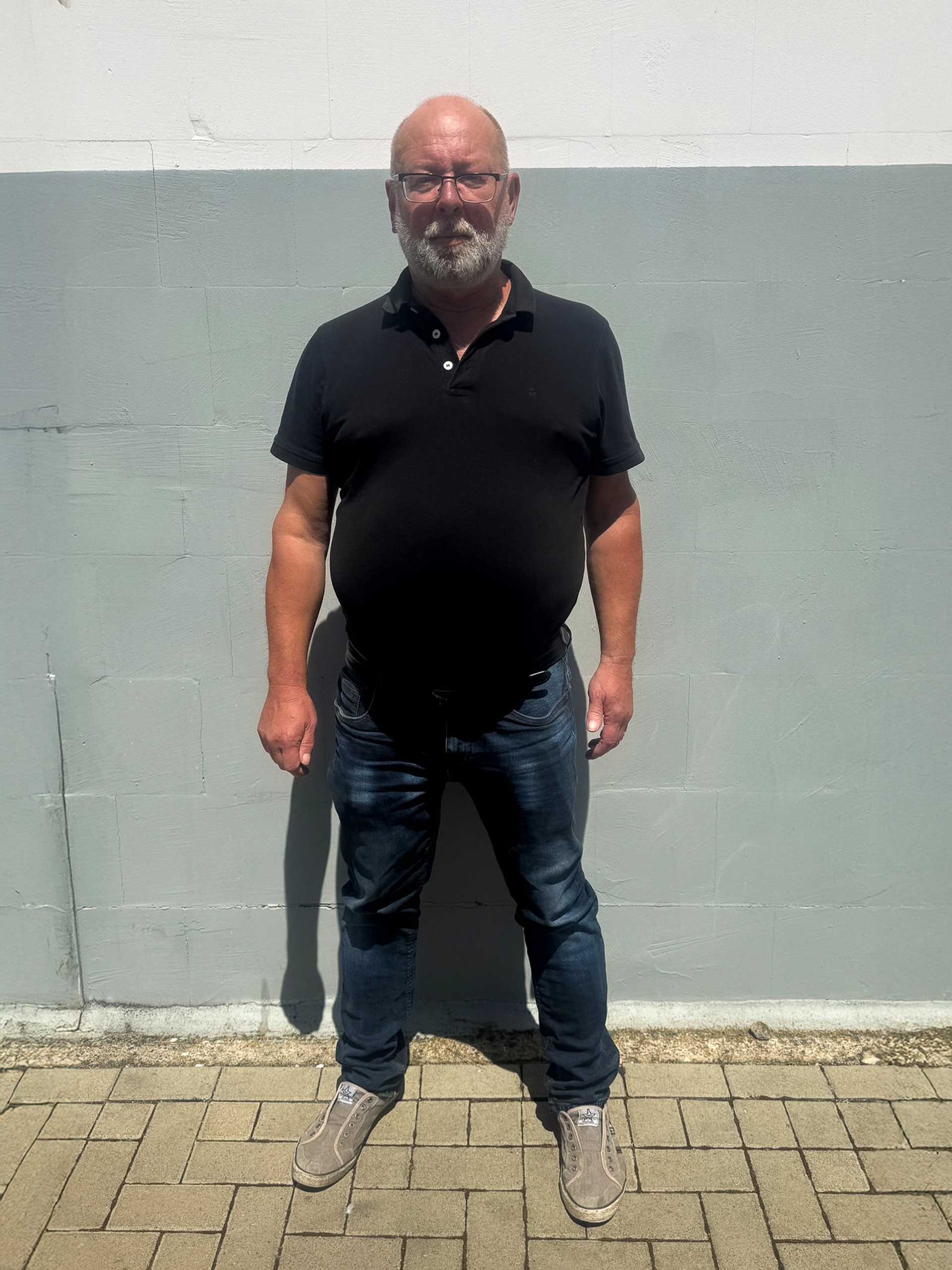Ralf Loschge steht mit schwarzem Polohemd, blauer Jeans und grauen Schuhen auf einer gepflasterten Fläche im Sonnenlicht vor einer zweifarbigen Wand - bereit für den Reifenservice oder Reifenwechsel bei Reifen Wolfenbüttel.