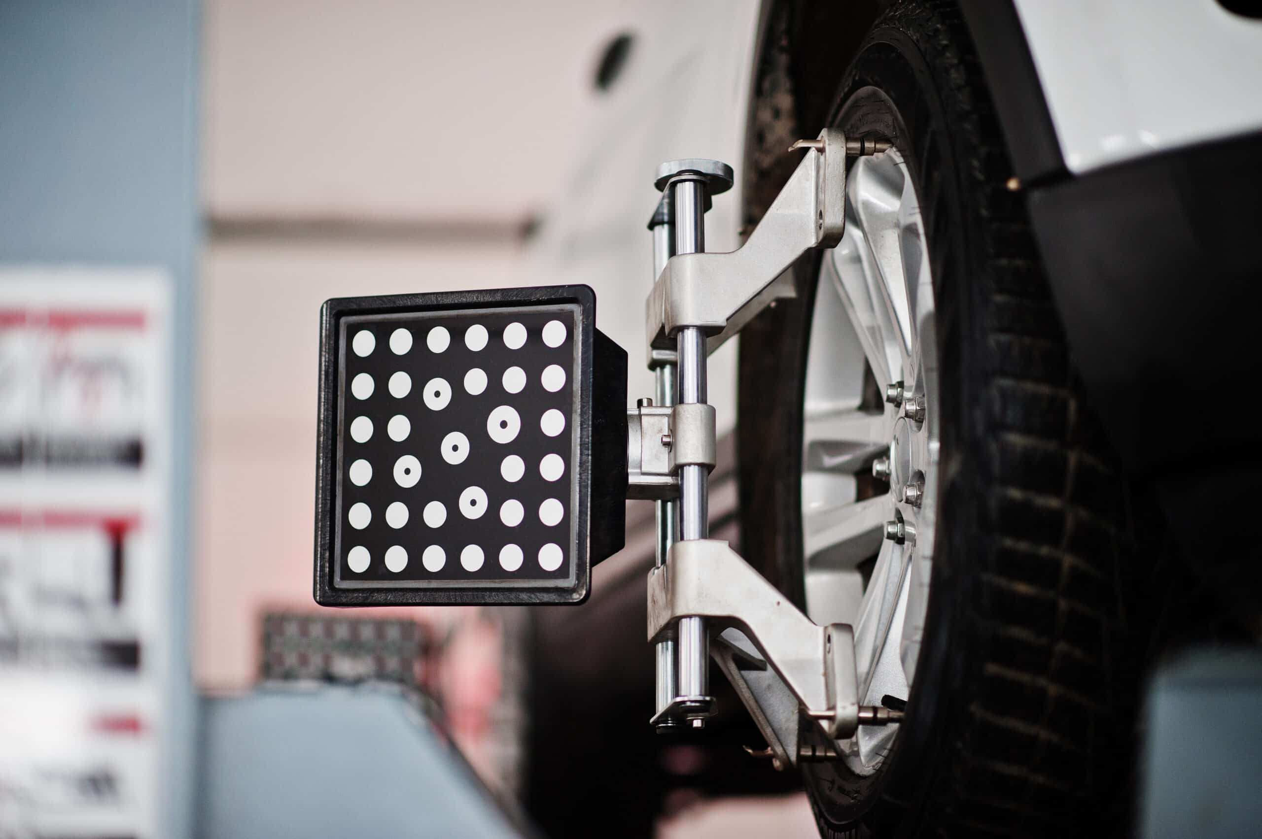 Nahaufnahme eines Autorads, an dem ein Ausrichtungswerkzeug angebracht ist, das eine quadratische Platte mit einem Gitter aus weißen Punkten zeigt. Der Hintergrund ist leicht verschwommen, was an eine Kfz-Werkstatt erinnert.
