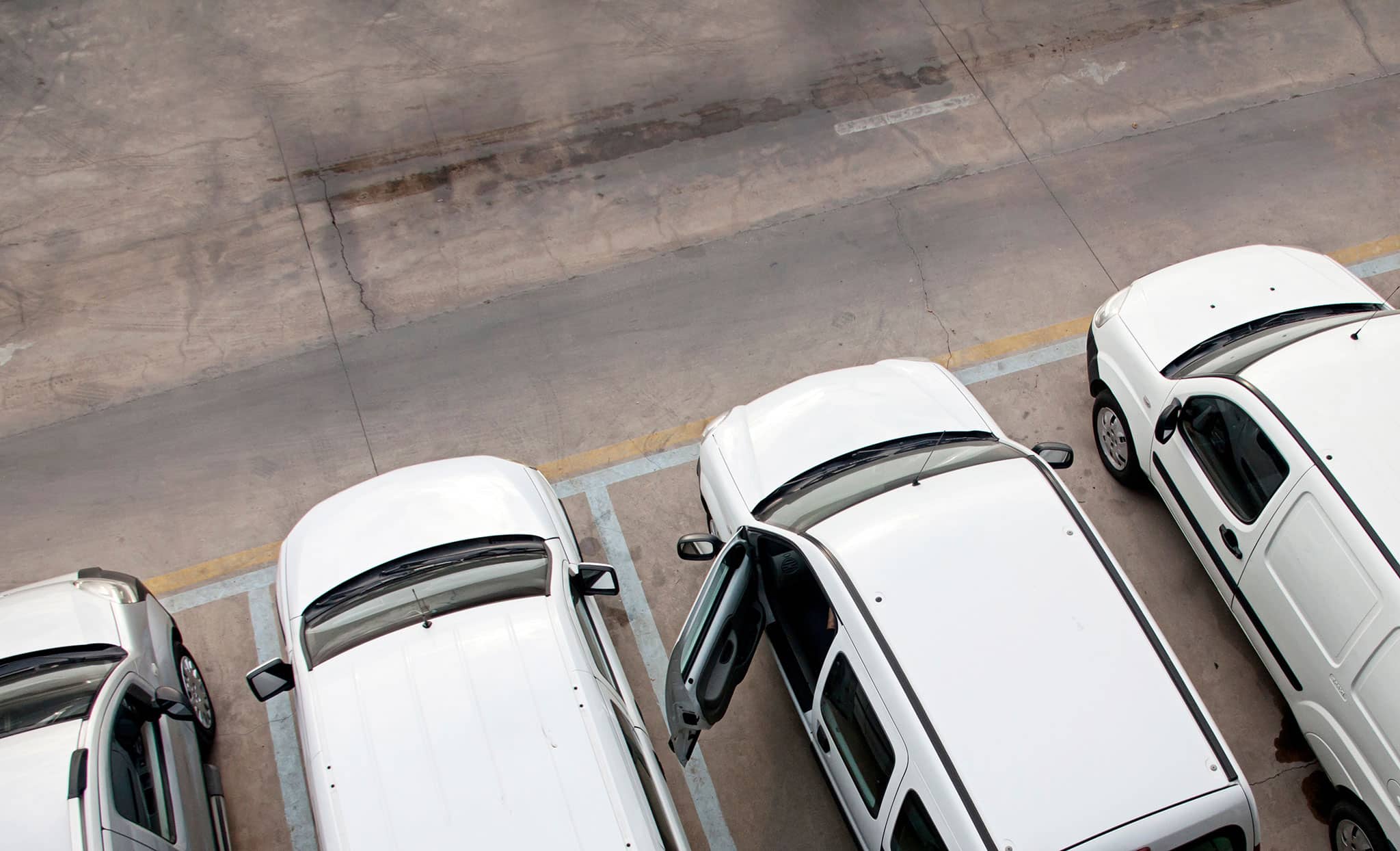 Draufsicht auf einen Parkplatz mit weißen Autos, die auf ausgewiesenen Plätzen geparkt sind; bei einem Auto ist die Fahrertür offen, die anderen sind mit geschlossenen Türen geparkt. Der Bürgersteig scheint sauber und leer zu sein.