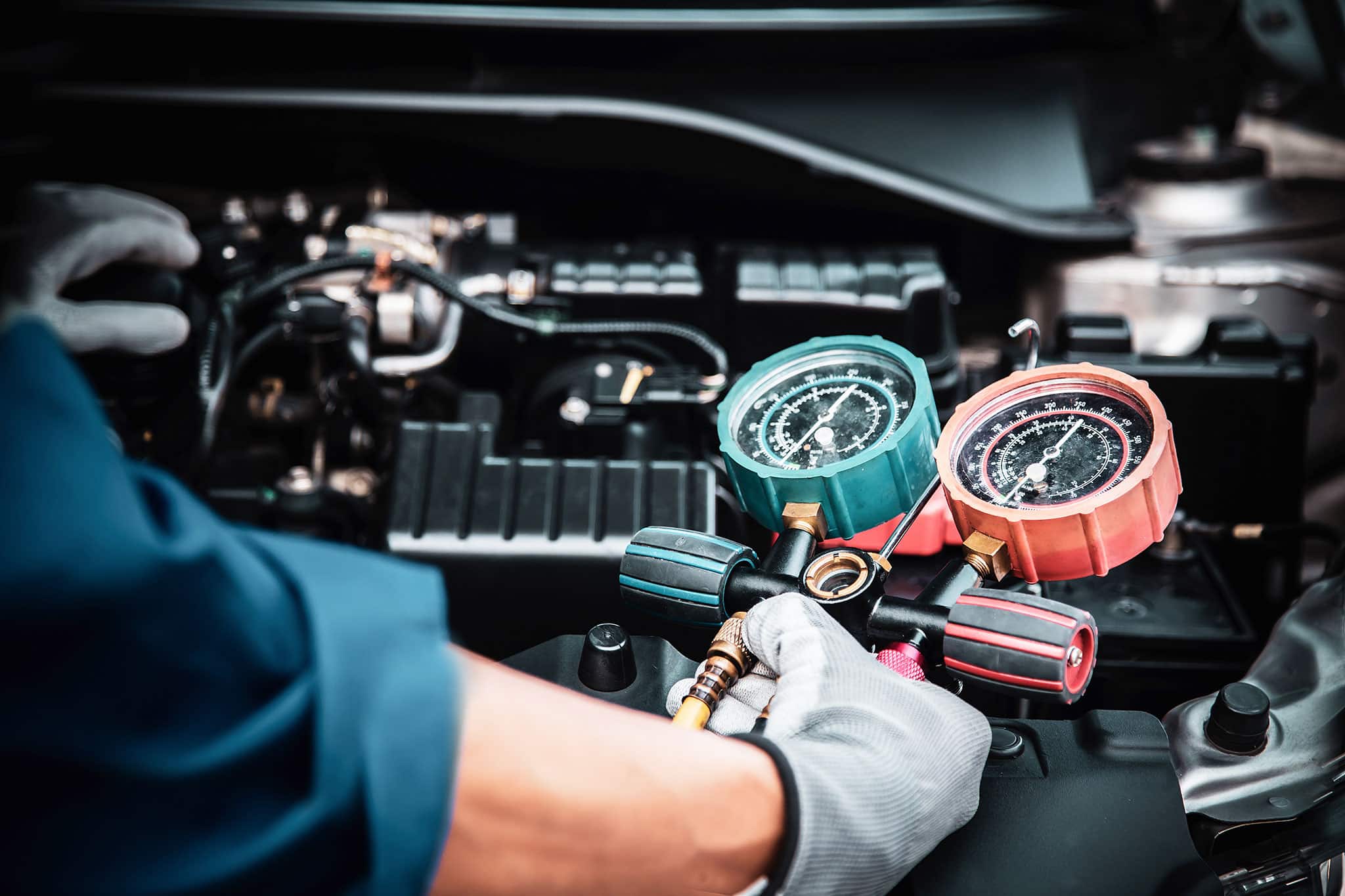 Eine Person mit Handschuhen benutzt ein Verteilermessgerät, um eine Klimaanlage unter der Motorhaube eines Fahrzeugs zu überprüfen oder zu warten. Die Anzeigen haben rote und blaue Ziffernblätter, und verschiedene Motorkomponenten sind sichtbar.