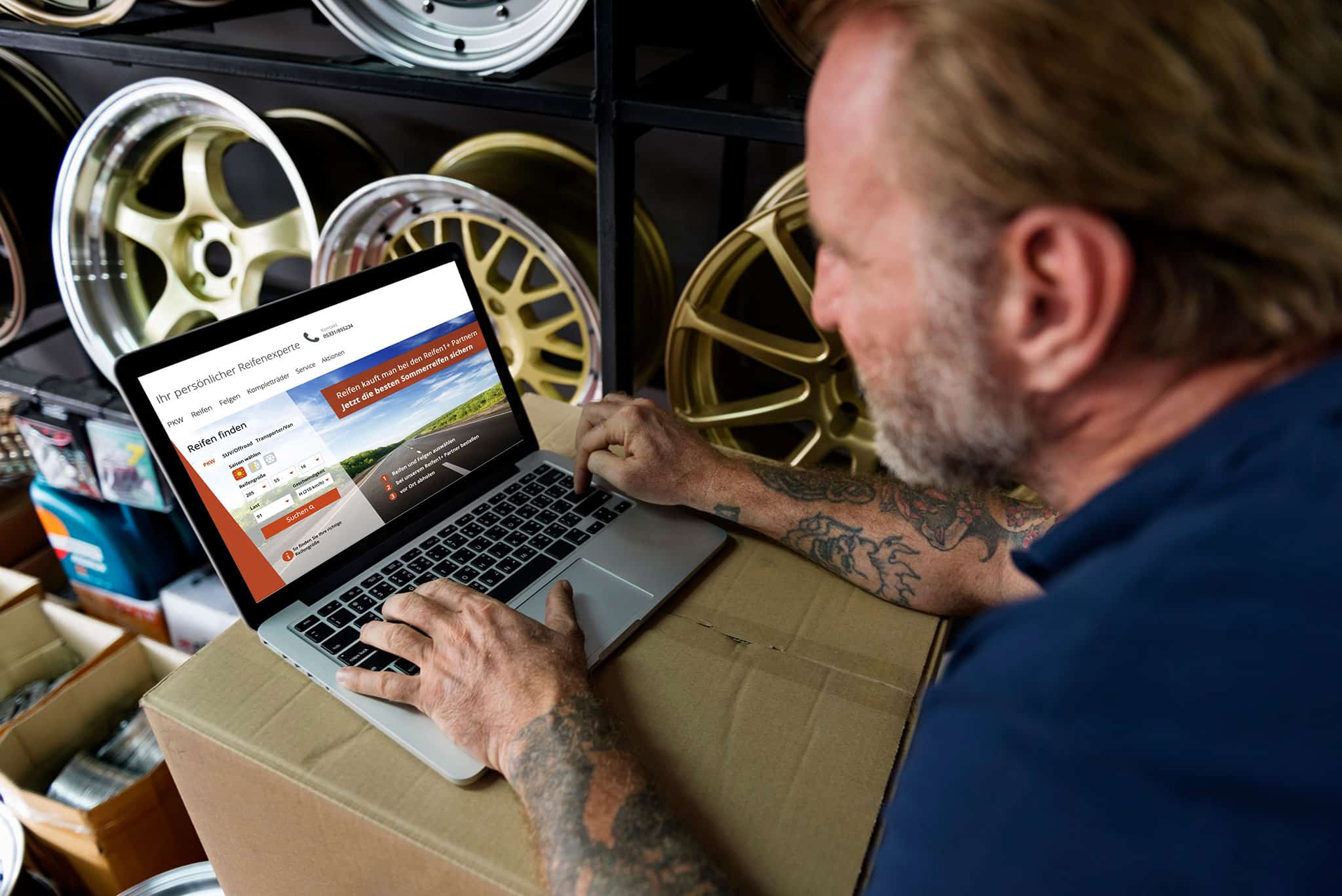 Eine Person mit Tattoos benutzt einen Laptop, der auf einem Karton in einer Garage oder Werkstatt mit Autofelgen auf Regalen steht. Auf dem Bildschirm des Laptops wird eine Website über Reifen oder Autos angezeigt.