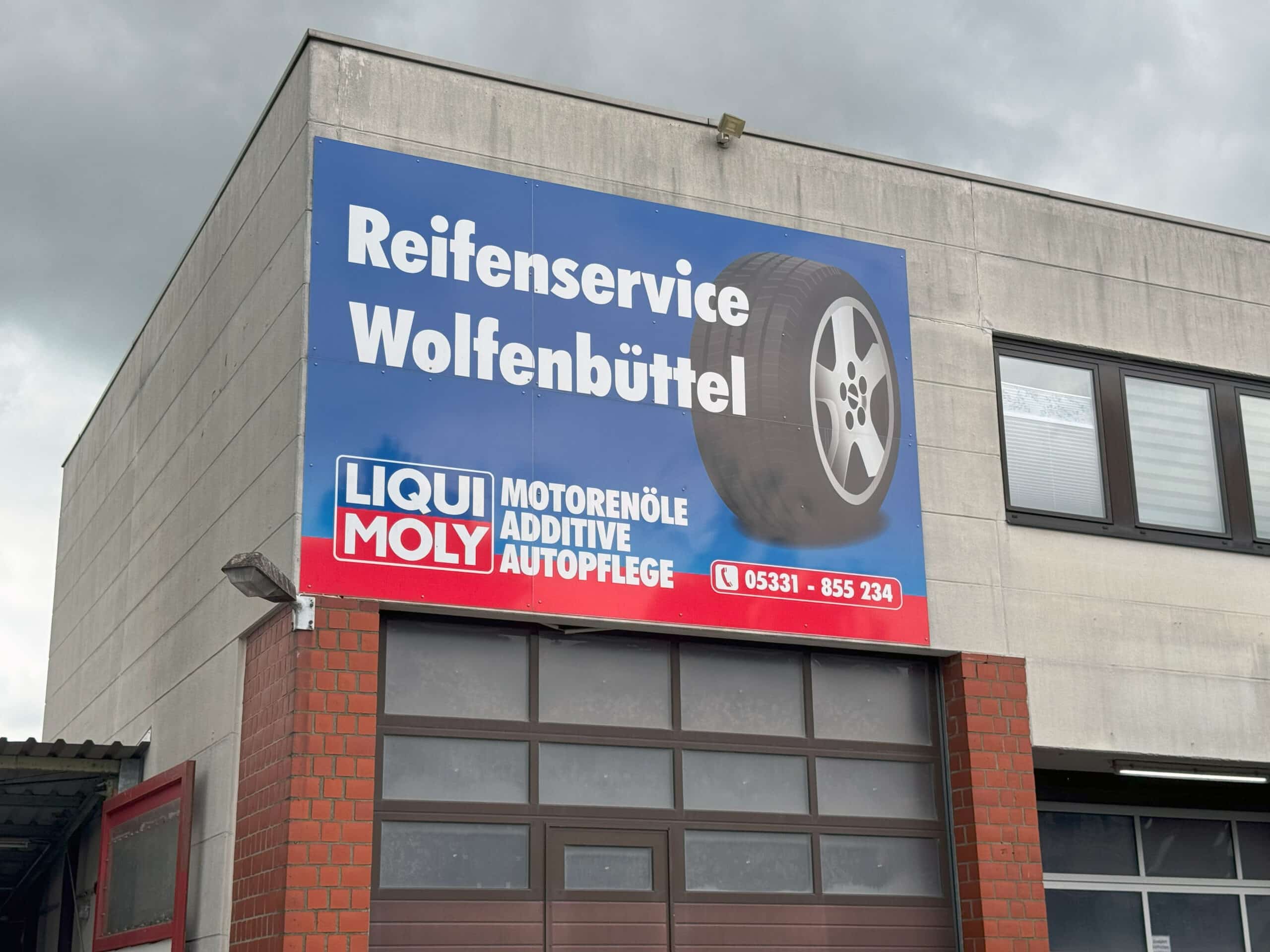 Auf einem grauen Gebäude ist ein Schild mit der Aufschrift "Reifenservice Wolfenbüttel" zu sehen, auf dem ein Autoreifen abgebildet ist, der auf einen Reifenwechsel hinweist. Logos für Liqui Moly und Kontaktdaten sind ebenfalls zu sehen. Der Himmel ist bewölkt.