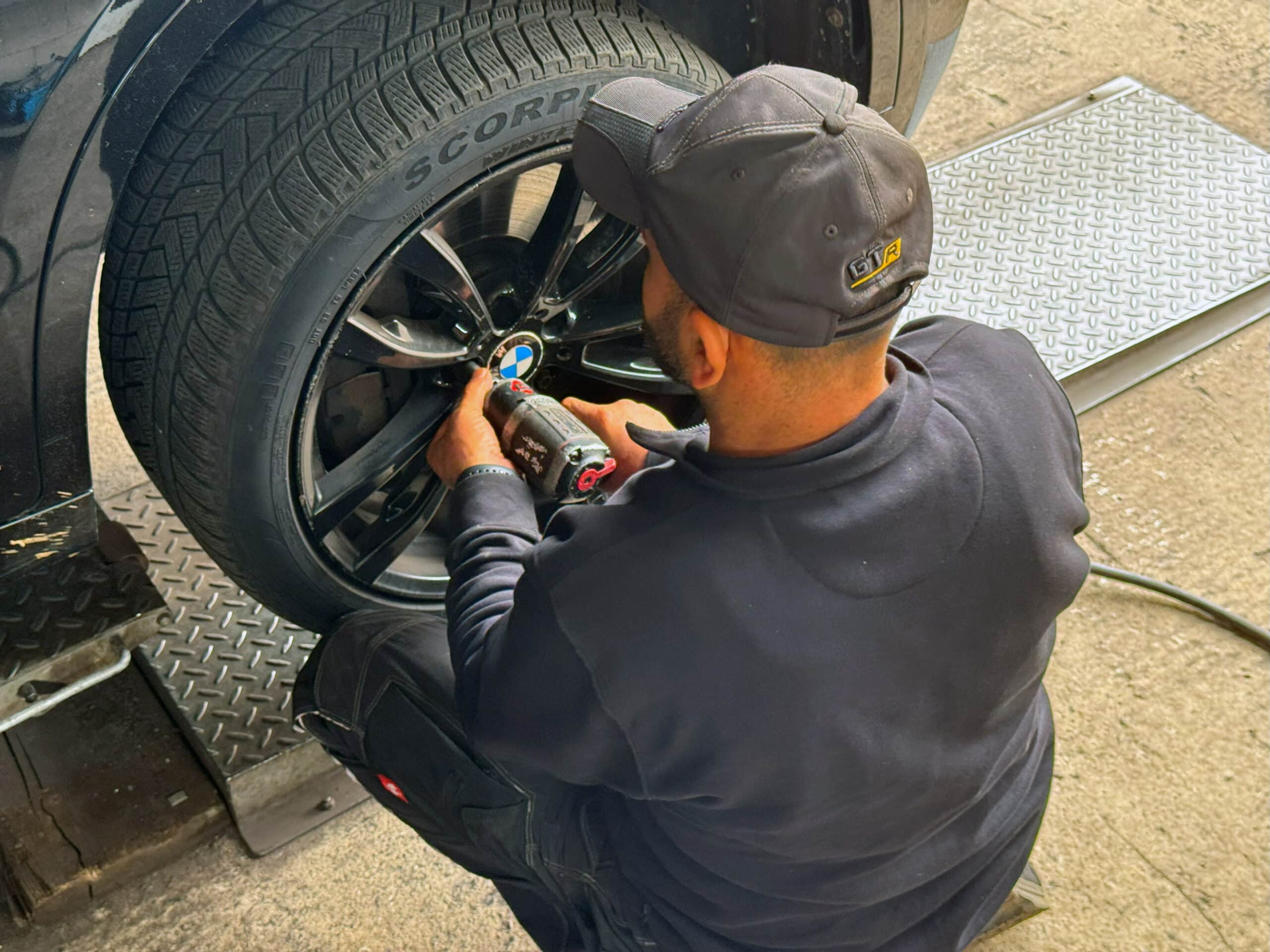Ein Mechaniker mit Mütze und dunkler Kleidung zieht während eines Reifenwechsels in einer Autowerkstatt mit einem Elektrowerkzeug die Radmuttern an einem schwarzen Autorad mit "Scorpion"-Reifen fest.