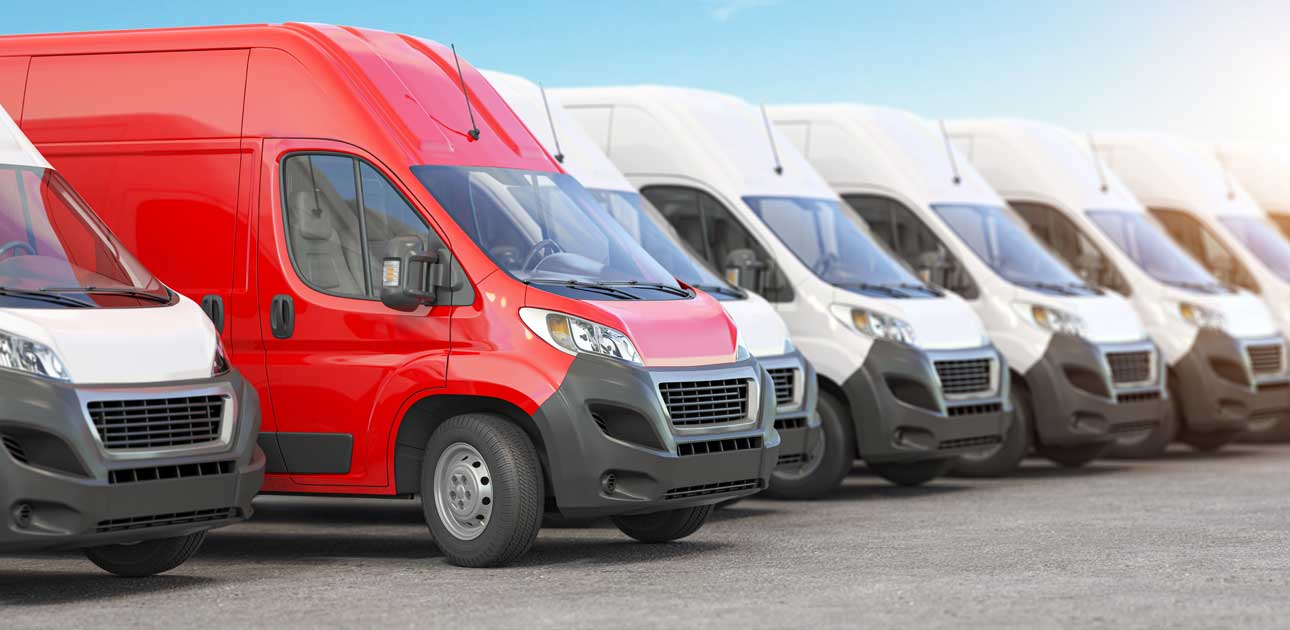 Eine Reihe von Lieferwagen parkt auf einem Parkplatz, ein roter Wagen sticht zwischen mehreren identischen weißen Lieferwagen hervor. Der klare Himmel bietet die perfekte Kulisse für einen schnellen Reifenwechsel oder Reifenservice bei Reifen Wolfenbüttel.