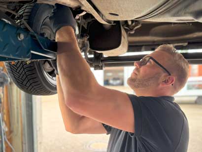 Ein Mann mit Brille und Handschuhen arbeitet in einer Werkstatt unter einem angehobenen Fahrzeug und konzentriert sich dabei auf das Fahrwerk und die Aufhängung des Wagens - möglicherweise als Vorbereitung für einen Reifenwechsel bei Reifen Wolfenbüttel.