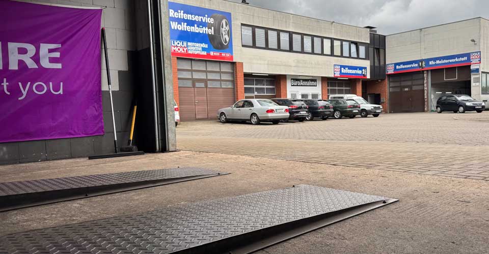 Außenansicht eines Autoservicezentrums mit mehreren Garagentoren, geparkten Autos und deutschen Schildern. Im Vordergrund ist eine metallene Autorampe zu sehen, während ein lila Banner auf den Reifenservice hinweist - Ihre Anlaufstelle für einen fachmännischen Reifenwechsel.