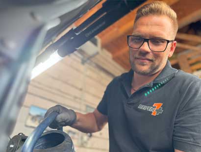 Ein Mann mit Brille und schwarzem Polohemd gießt in einer Werkstatt Flüssigkeit aus einem blauen Behälter in einen Automotor und verdeutlicht damit den professionellen Reifenservice bei Reifen Wolfenbüttel.