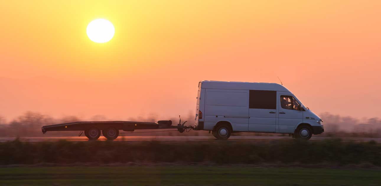 Ein weißer Lieferwagen mit Pritschenanhänger fährt bei Sonnenuntergang eine Straße entlang, die Sonne steht tief am Himmel und im Hintergrund ist ein Feld zu sehen - perfekt für Reifenservice vor Ort oder schnellen Reifenwechsel unterwegs.