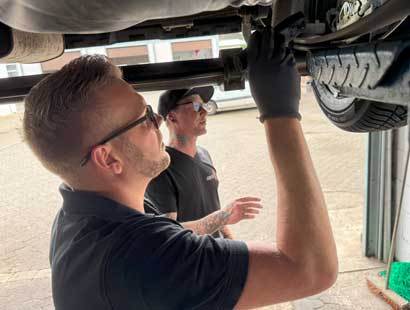 Zwei Männer stehen neben einem Fahrzeug auf einer Hebebühne beim Reifenservice Wolfenbüttel. Der eine, der Handschuhe und eine Schutzbrille trägt, arbeitet an der Unterseite des Fahrzeugs - möglicherweise beim Reifenwechsel -, während der andere zuschaut. Beide sind mit schwarzen Hemden bekleidet.