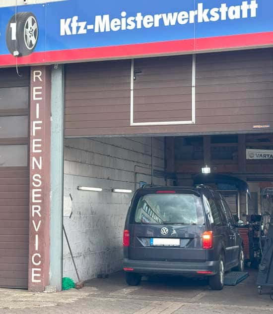 Ein schwarzer VW-Bulli steht in einer Werkstatt mit den Aufschriften "Kfz-Meisterwerkstatt" und "Reifenservice", was darauf hindeutet, dass es sich um eine Autowerkstatt handelt, die Reifendienste wie Reifenwechsel anbietet.