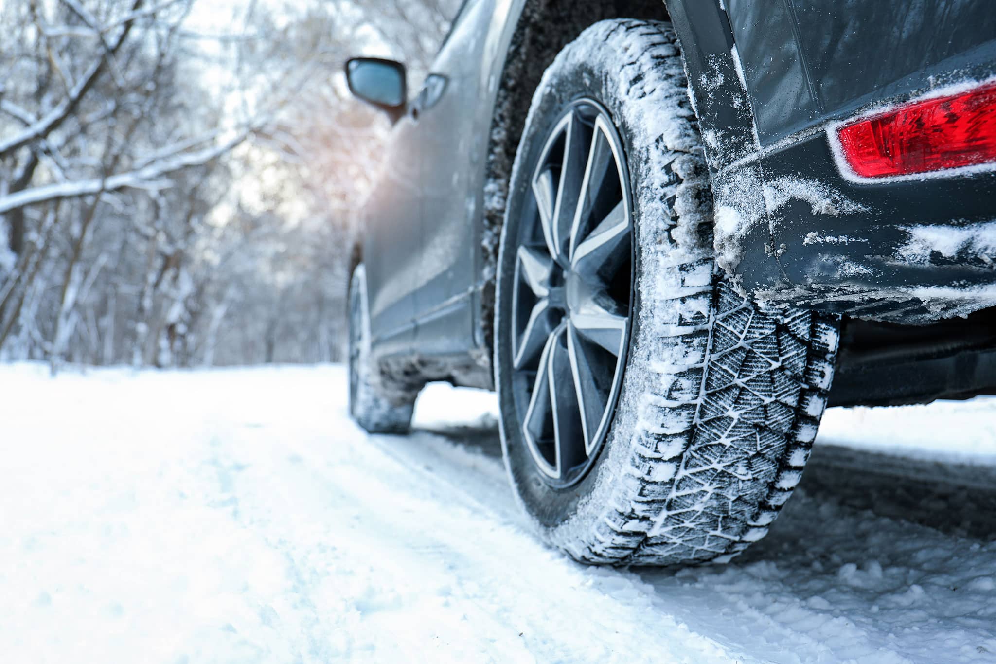 Nahaufnahme des Hinterrads eines Autos mit Winterreifen, das auf einer verschneiten Straße fährt. Schnee bedeckt den Boden und die Bäume auf beiden Seiten der Straße. Das Auto hat sichtbaren Schnee und Eis um das Rad und die unteren Teile.